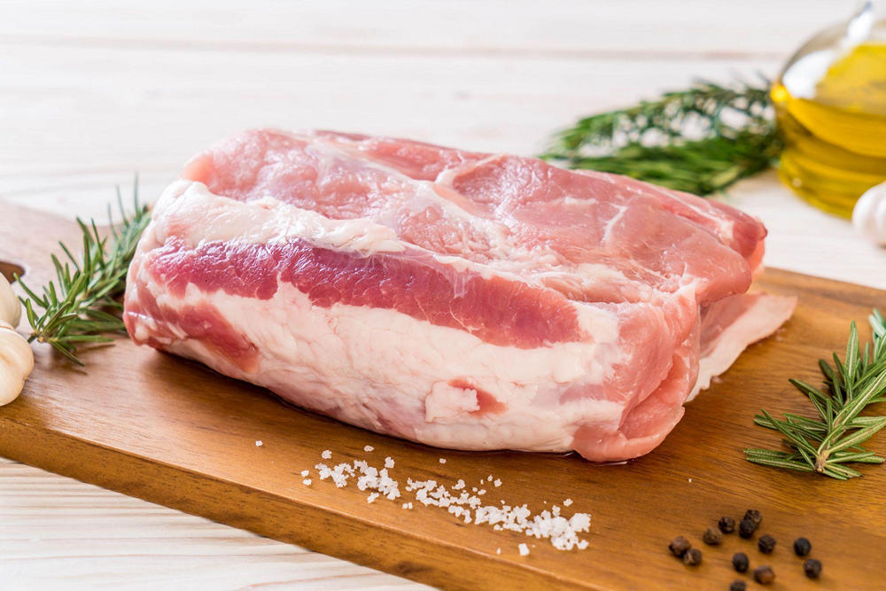 На деревянной доске кусок сырой свинины, рядом крупицы соли, горошины перца и веточки зелени