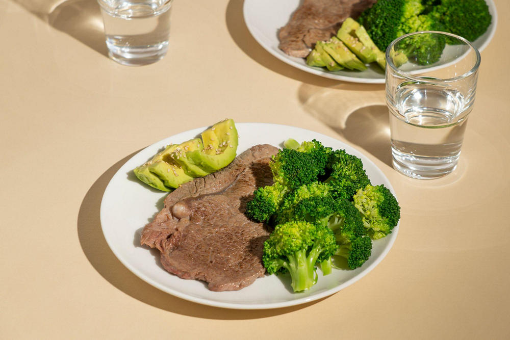 На столе тарелка с отварной говядиной, авокадо и брокколи. Рядом стакан воды.