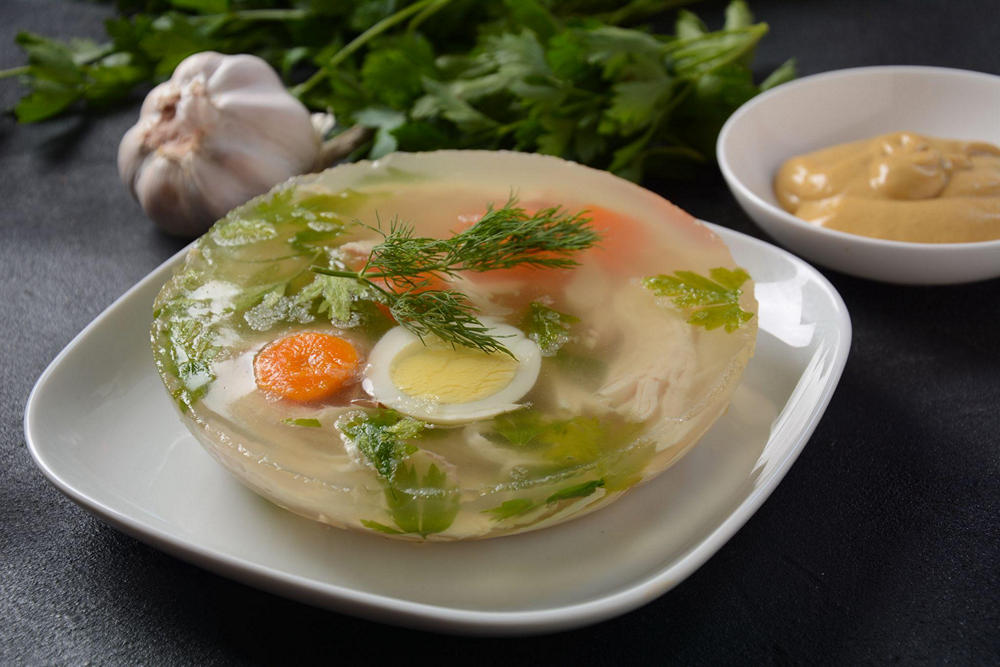 На столе тарелка с холодцом круглой формы с яйцом, зеленью и морковкой