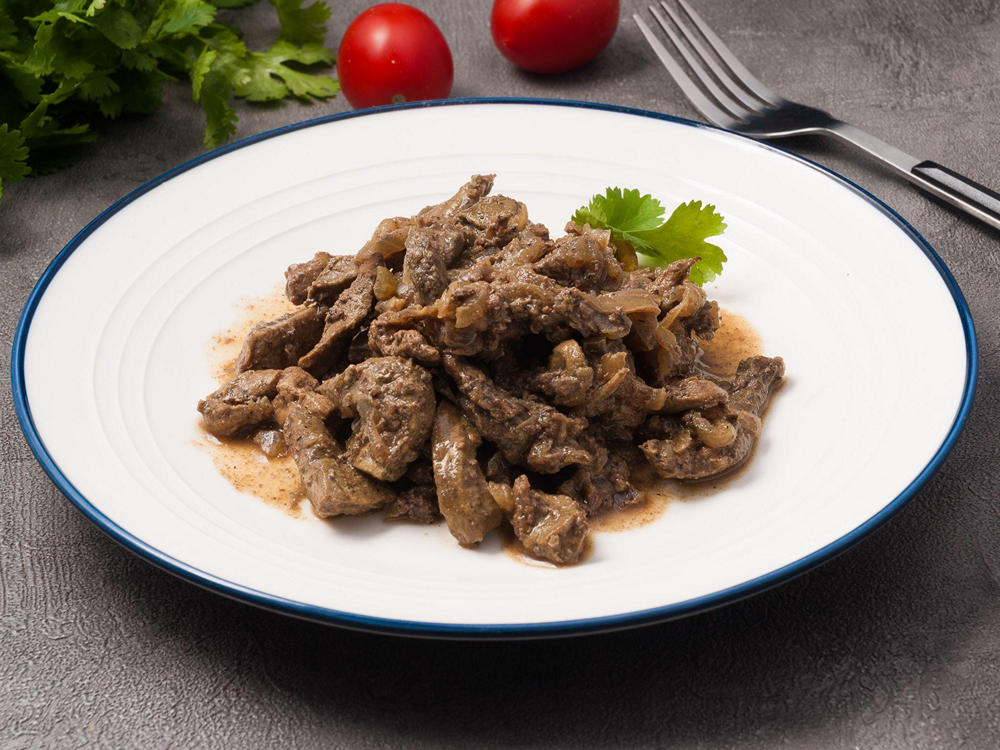 На столе тарелка с готовым блюдом: бефстроганов из говядины, украшенный зеленью петрушки