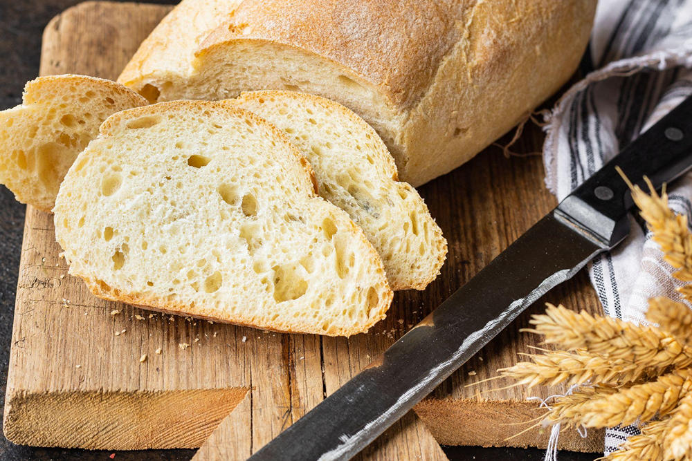 На деревянной доске булка белого хлеба и нарезанные куски, рядом лежит нож для нарезки и колосья