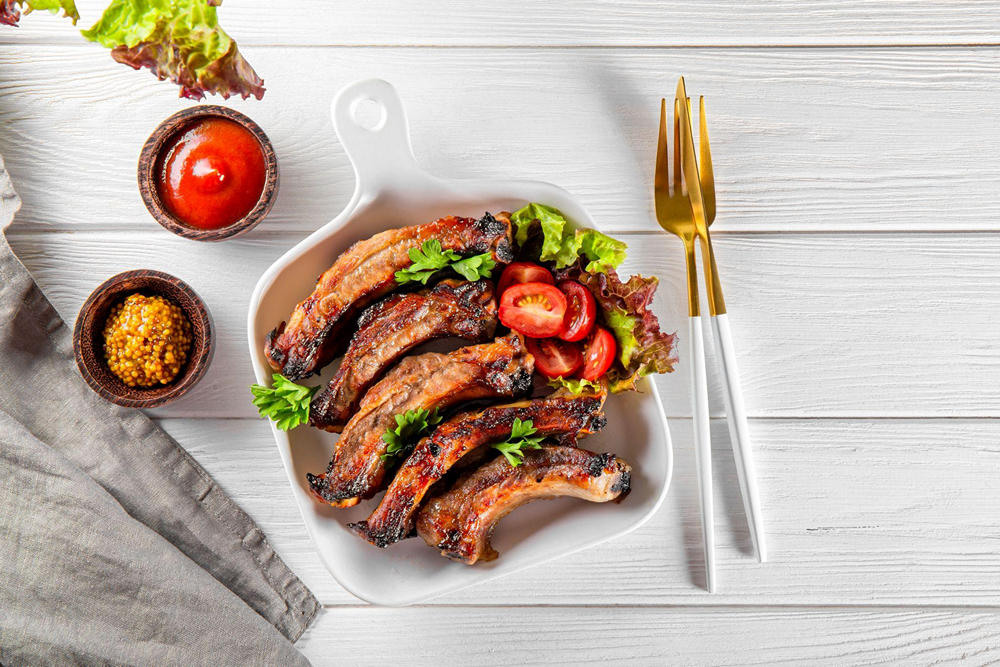 На столе тарелка с свиными ребрами барбекю, украшенными зеленым салатом и помидорами. Рядом столовые приборы, соус и горчица