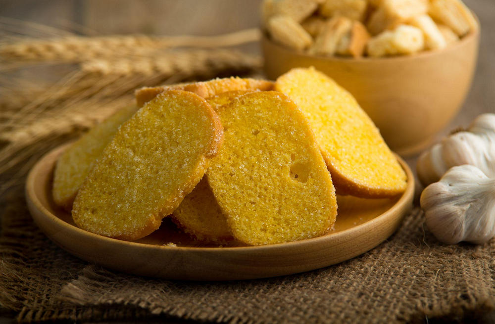 На столе деревянная тарелка с кусками желтого кукурузного хлеба, рядом головка чеснока