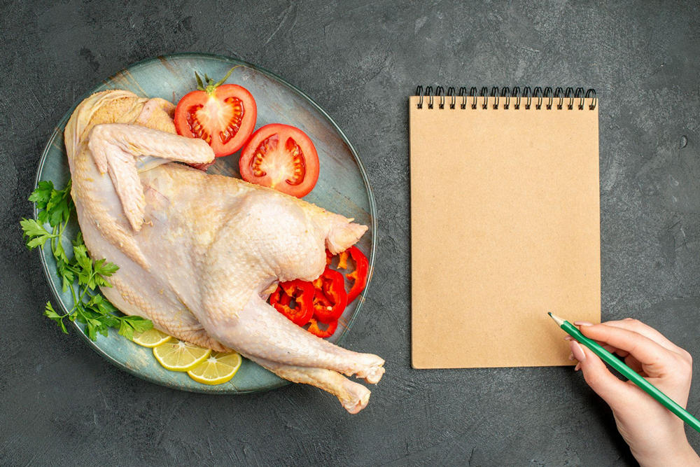 Человек составляет список учета калорий для диеты, держа карандаш над блокнотом. Рядом на столе стоит тарелка с индейкой, дольками лимона, зеленью и овощами: помидорами и перцами