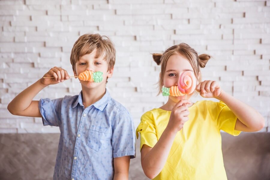 Мальчик и девочка, играют вместе в помещении. Каждый из них подносит ко рту леденцы на палочках, и позирует для фото на фоне белой кирпичной стены