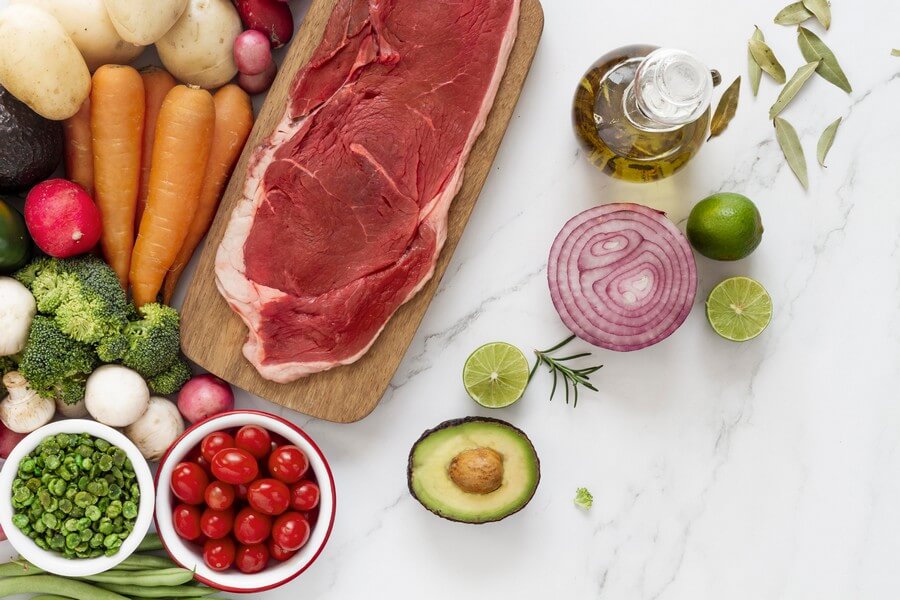 Фотография разнообразных свежих овощей и зелени, таких как брокколи, морковь, картофель, грибы, зеленые бобы, томаты, болгарский перец, лук, чеснок, авокадо, кинза и лайм, а также кусок мяса на деревянной разделочной доске на светлом фоне