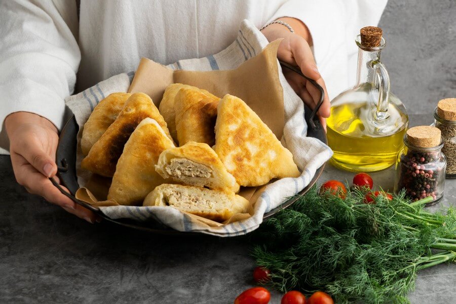 Фотография с пирогами на металлическом подносе, рядом с которым расположены зелень, томаты, маслины и бутылка масла, все это расположено на темной поверхности с элементами декора