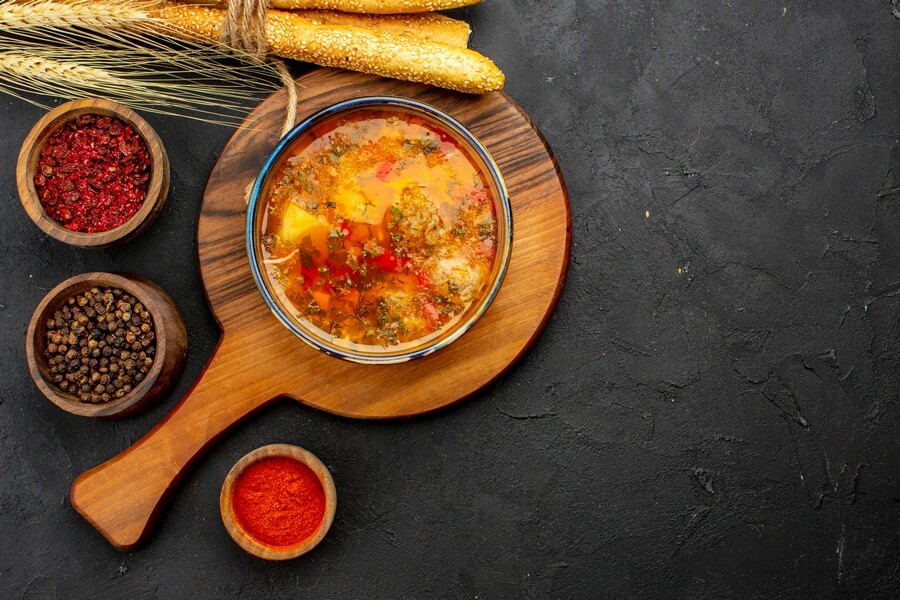 Тарелка с щами, украшенным свежей зеленью, рядом с различными специями и травами, солонкой и перечницей