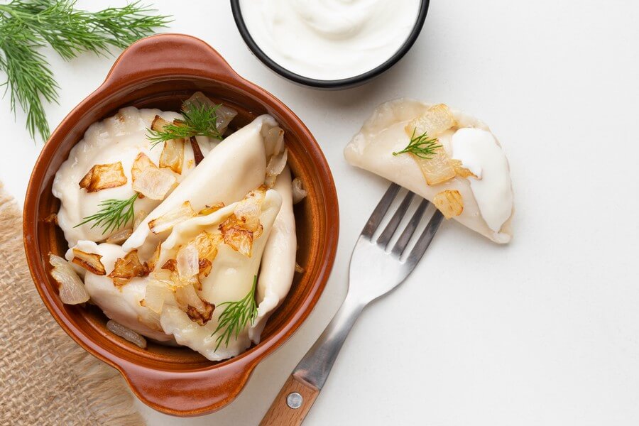 Тарелка с варениками, украшенными зеленью и посыпанными жареными луковыми кольцами, рядом находятся столовые приборы